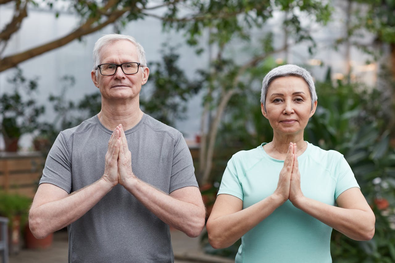 about-01 Senior couple performing yoga poses in a peaceful greenhouse setting.