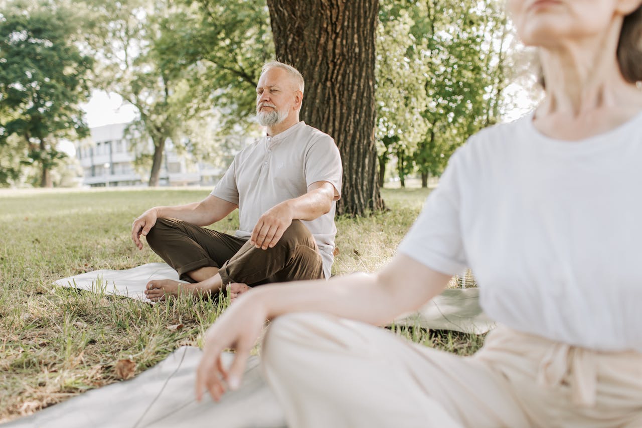 services-01 Elderly man and woman meditating outdoors on yoga mats in a peaceful park setting.