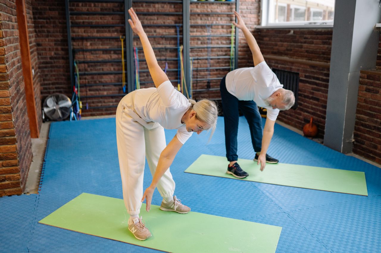 hero-img-01 Active senior couple performing stretching exercises on mats in a modern fitness gym.